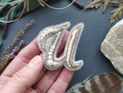A hand holding a decorative letter 'U' made of beads, pearls, and other embellishments against a dark background with various natural elements like leaves and feathers.