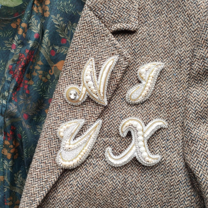 a close-up view of a tweed jacket with four decorative letter brooches attached to it.