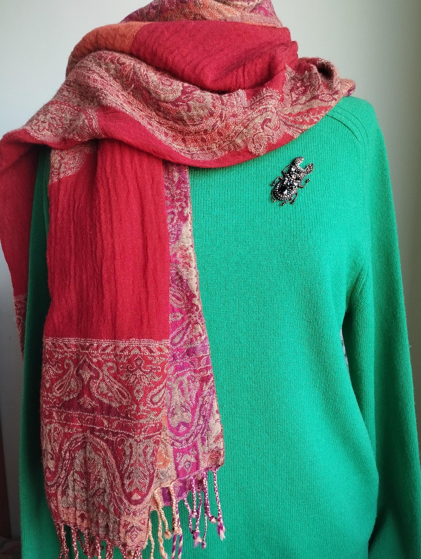 A mannequin is wearing a green sweater with a red and pink paisley-patterned scarf draped over it. The mannequin also has a brooch on its left shoulder.
