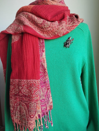A mannequin is wearing a green sweater with a red and pink paisley-patterned scarf draped over it. The mannequin also has a brooch on its left shoulder.