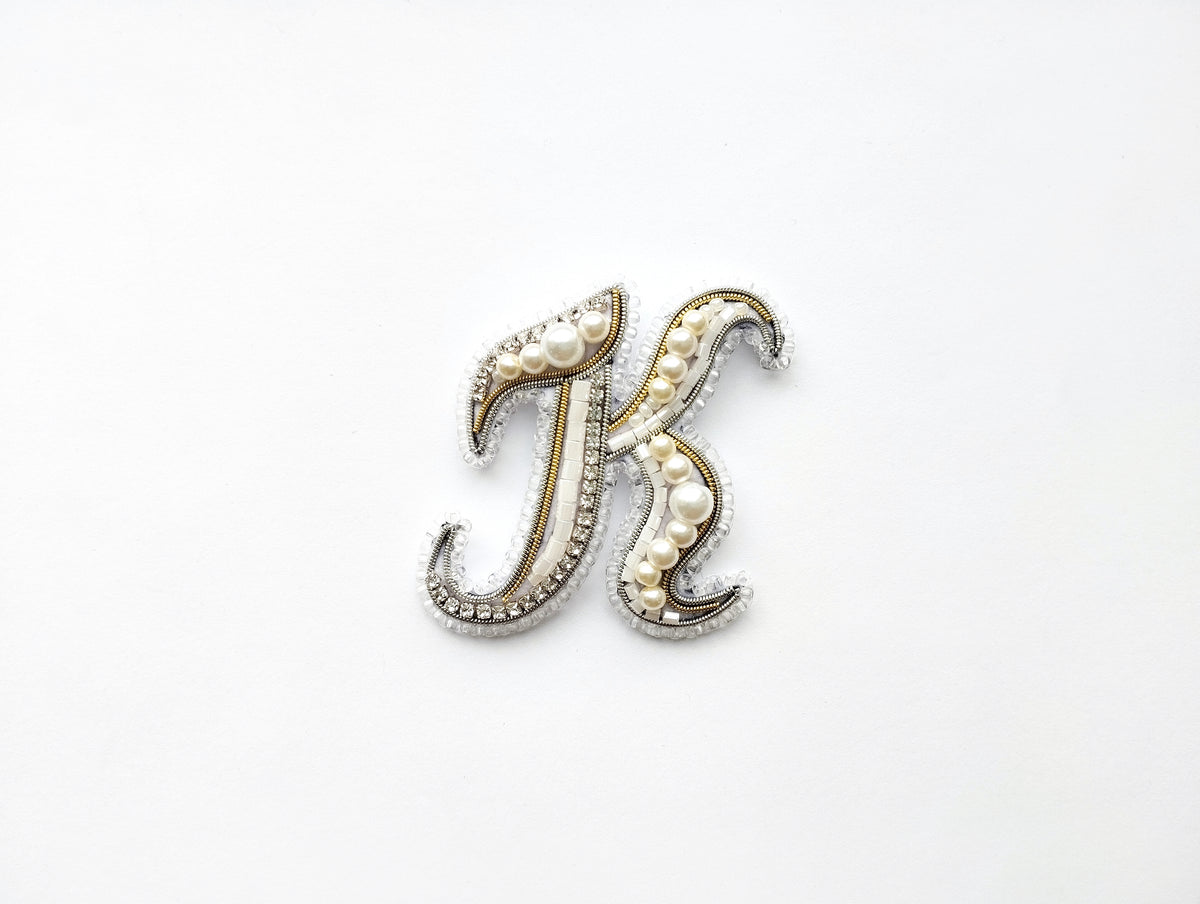 A decorative letter 'K' made of beads, pearls, and metallic elements, placed against a white background.