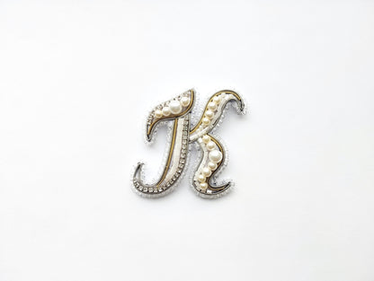 A decorative letter 'K' made of beads, pearls, and metallic elements, placed against a white background.