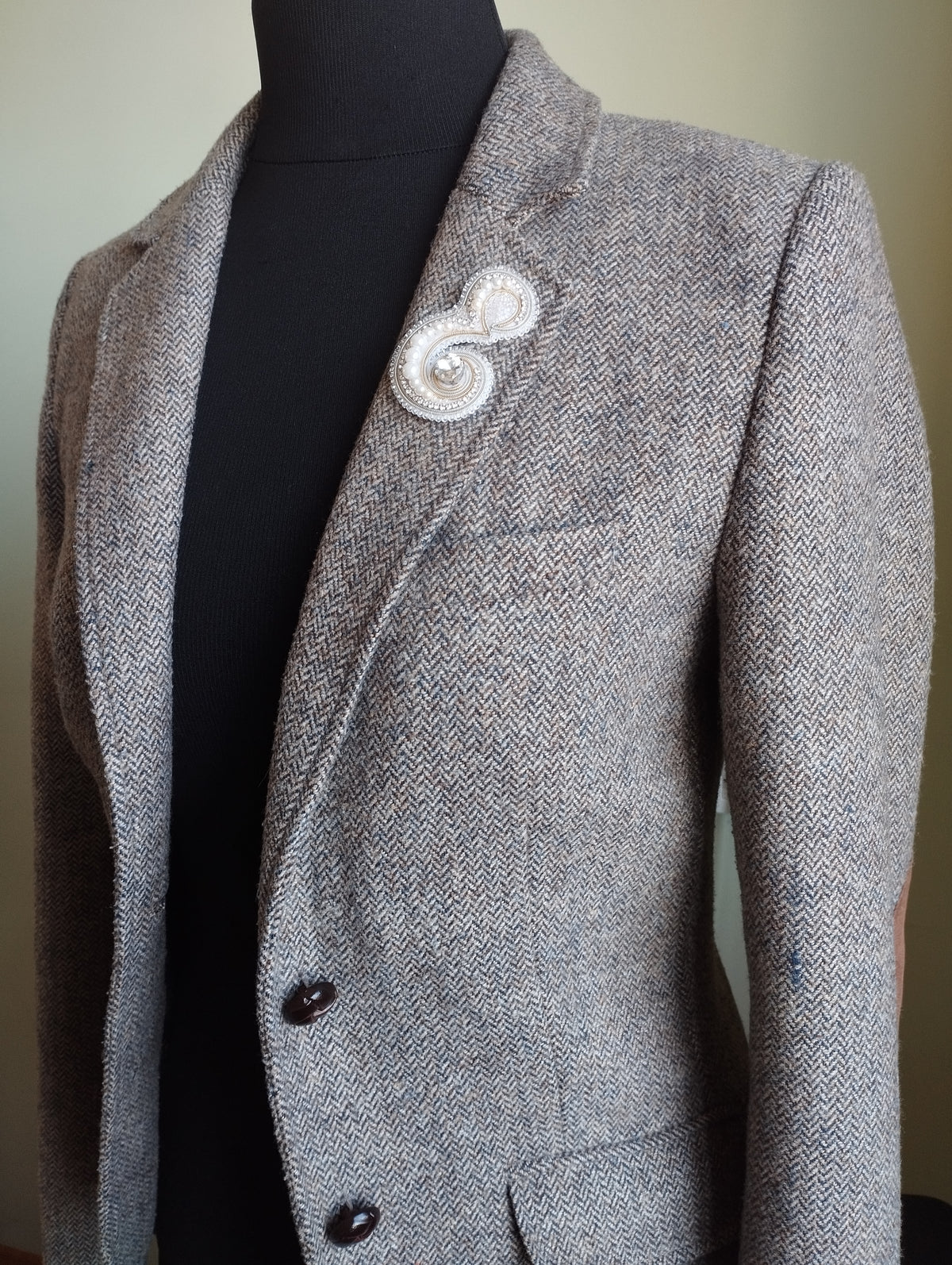 A gray tweed blazer with a white brooch on the left side of the chest.