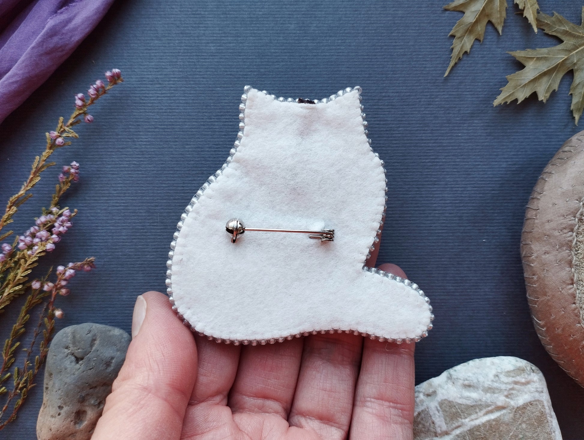 A hand holding a white felt cat pin against a dark background with various objects, including leaves and rocks.