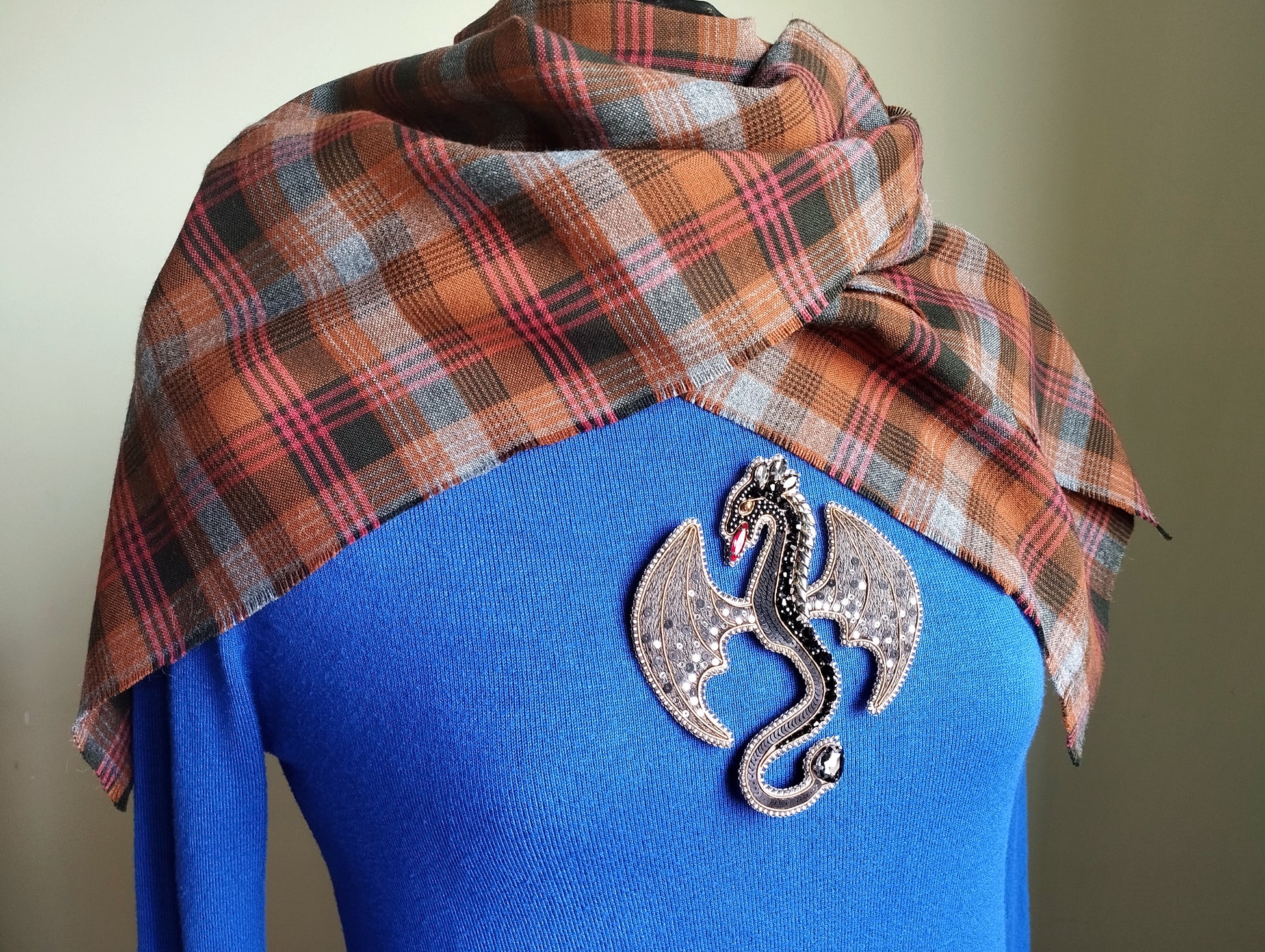 A blue sweater with a plaid scarf draped over it, and a decorative dragon brooch pinned to the front of the sweater.