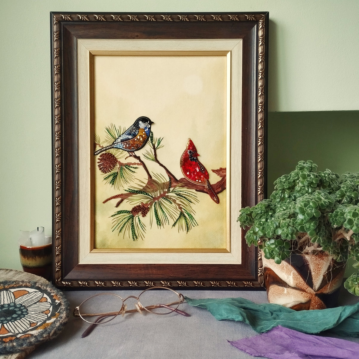 a framed painting of two birds perched on a branch, surrounded by pine cones and greenery, placed on a table next to a potted plant.