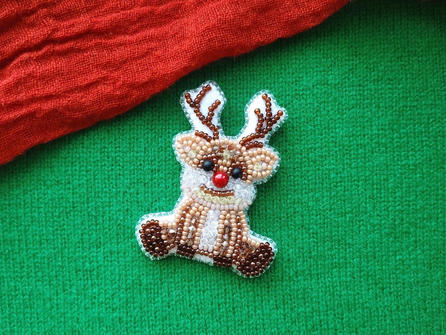 A small, beaded reindeer pin is displayed on a green fabric surface, with a red fabric item partially visible in the background.
