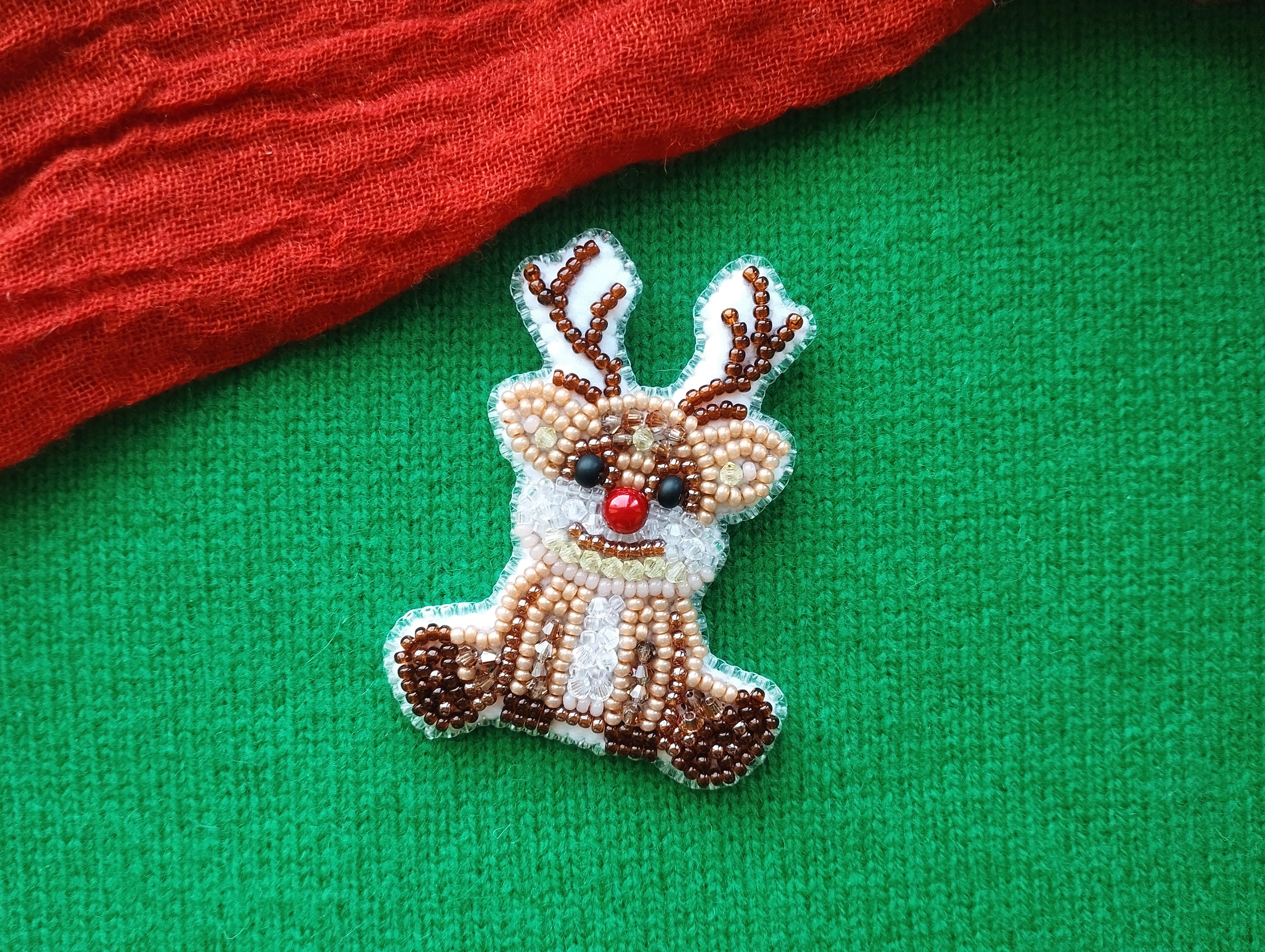 A small, beaded reindeer pin is displayed on a green fabric surface, with a red fabric item partially visible in the background.