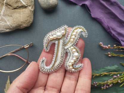 A hand holding a decorative letter "K" made of beads and pearls, surrounded by various objects such as a rock, a purple fabric, and some flowers.