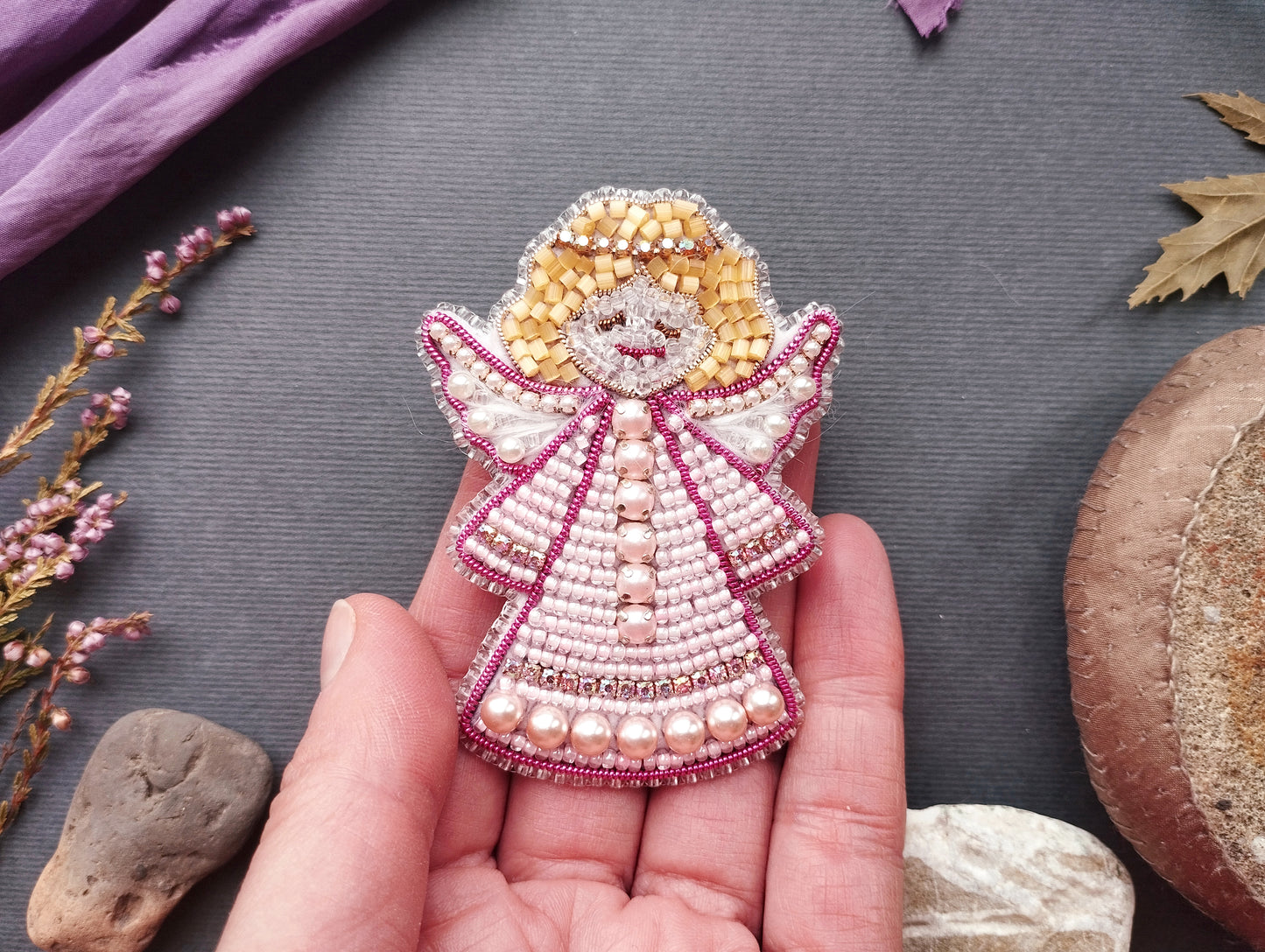 A hand holding a small, intricately beaded angel pin against a gray background.