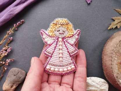 A hand holding a small, intricately beaded angel pin against a gray background.