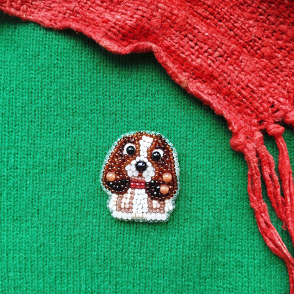 A small embroidered dog brooch is displayed on a green fabric, with a red fabric or material partially visible on the right side.