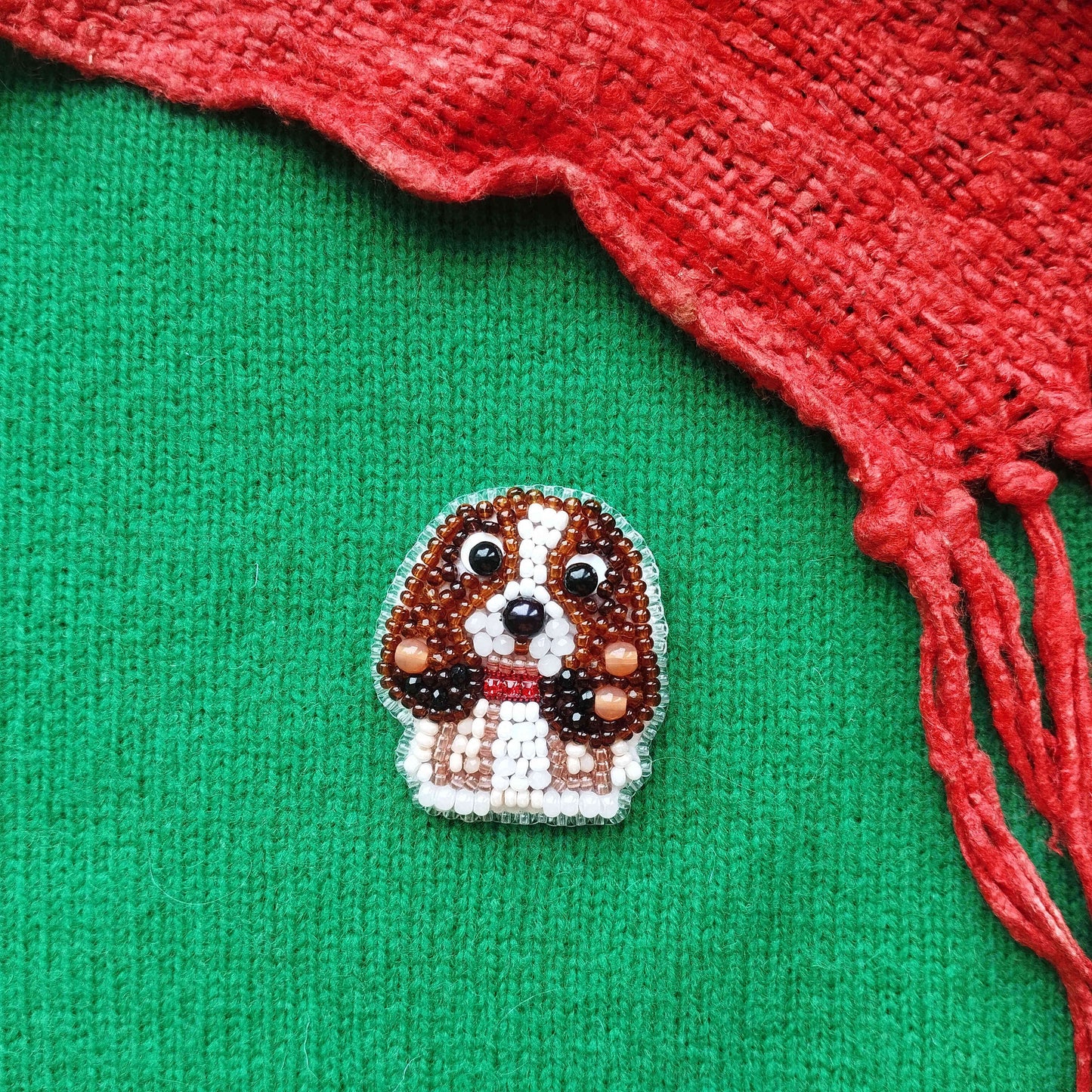 A small embroidered dog brooch is displayed on a green fabric, with a red fabric or material partially visible on the right side.