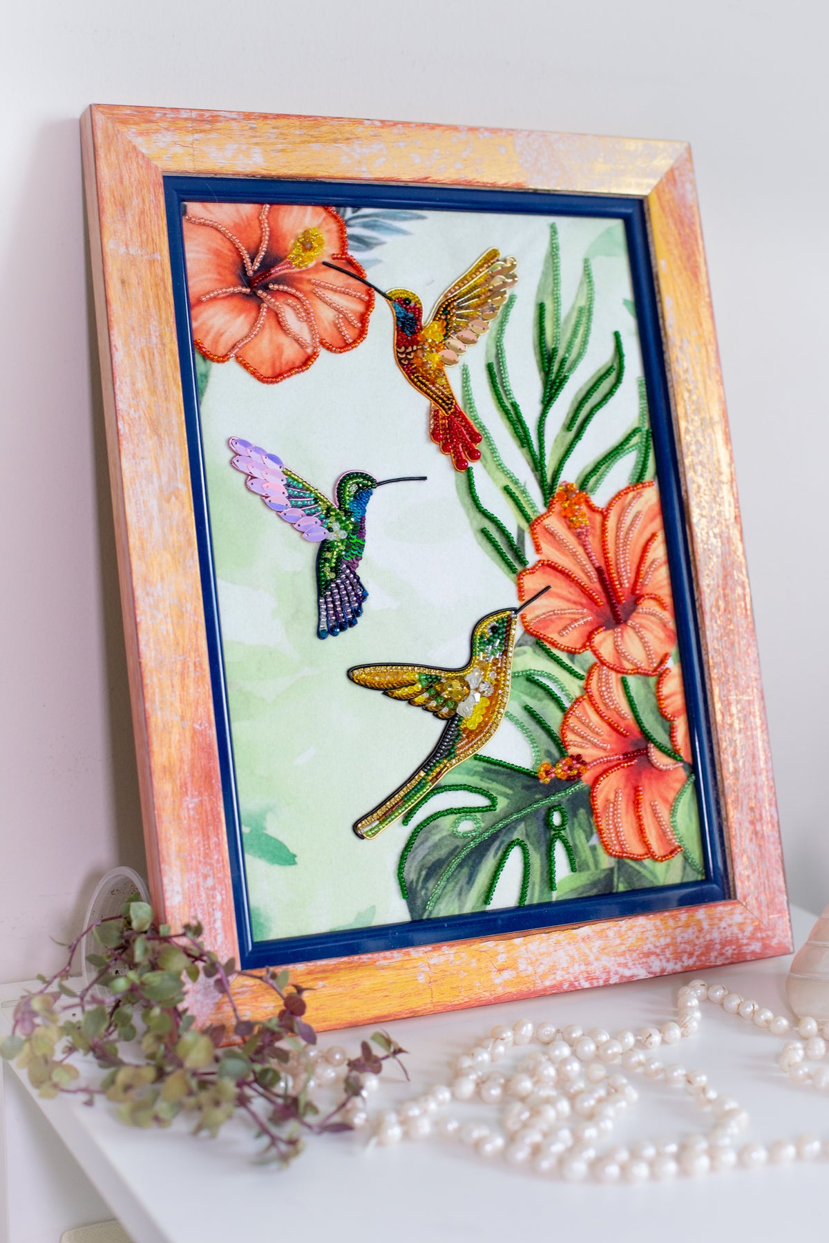 a framed painting of three colorful hummingbirds hovering over vibrant flowers, with a lush green background.