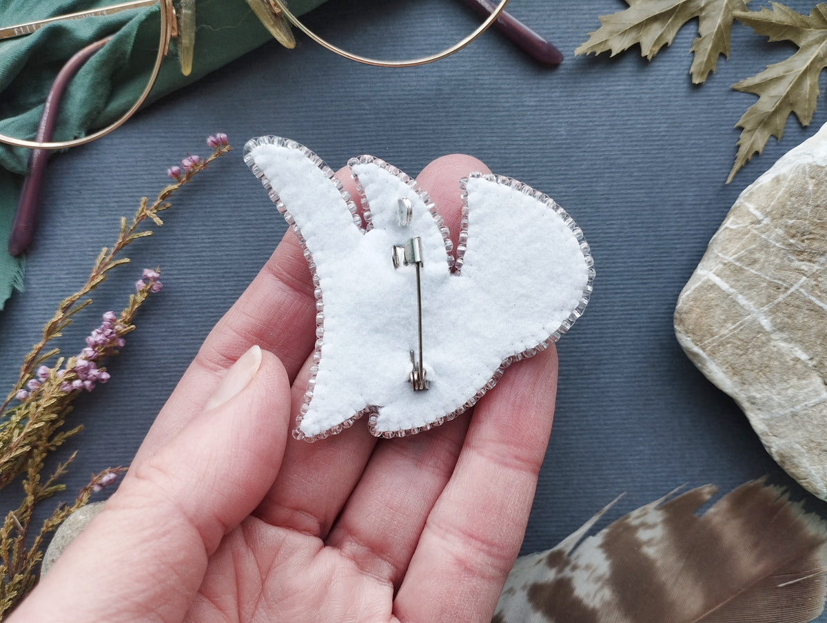 A hand holding a white felt pin or badge with a silver pin attached, surrounded by various natural elements such as leaves, feathers, and rocks.