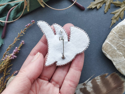 A hand holding a white felt pin or badge with a silver pin attached, surrounded by various natural elements such as leaves, feathers, and rocks.