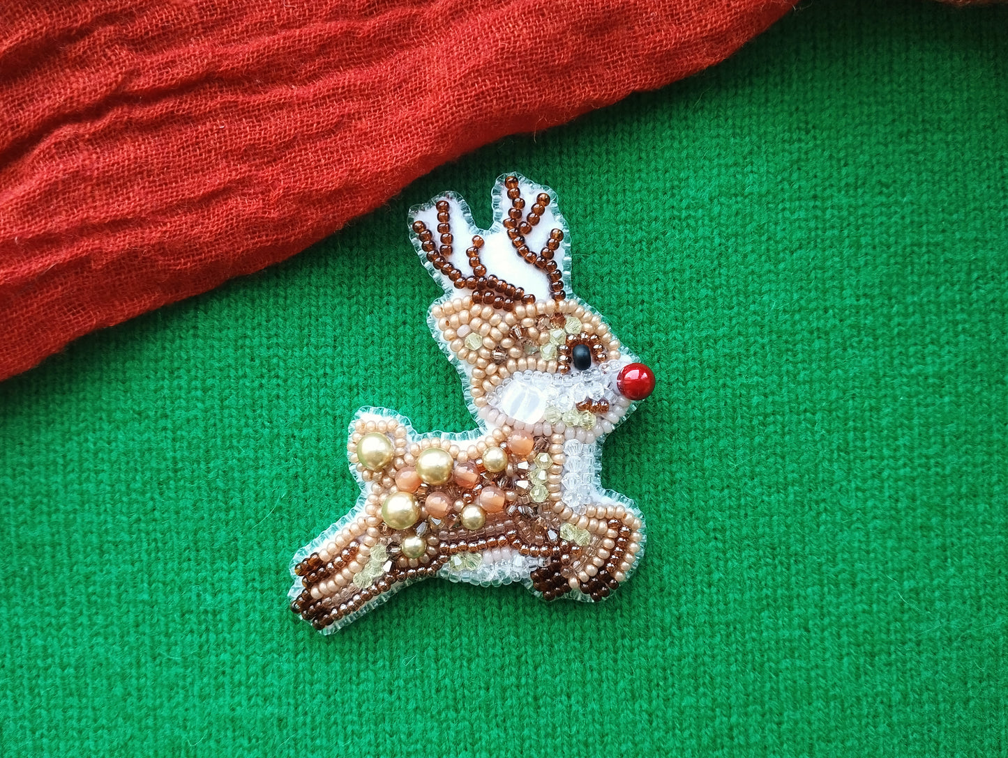 A beaded reindeer pin is displayed on a green fabric, with a red fabric piece partially visible to the left.