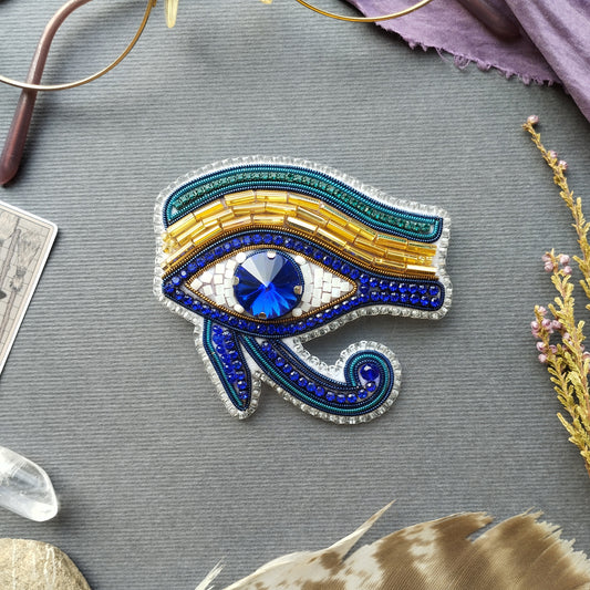 a close-up of a decorative eye brooch with blue and gold beads, placed on a gray surface.