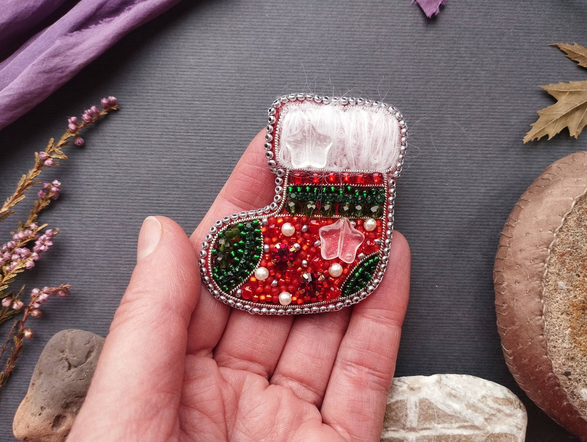A hand holding a small, intricately beaded Christmas stocking brooch against a dark background.