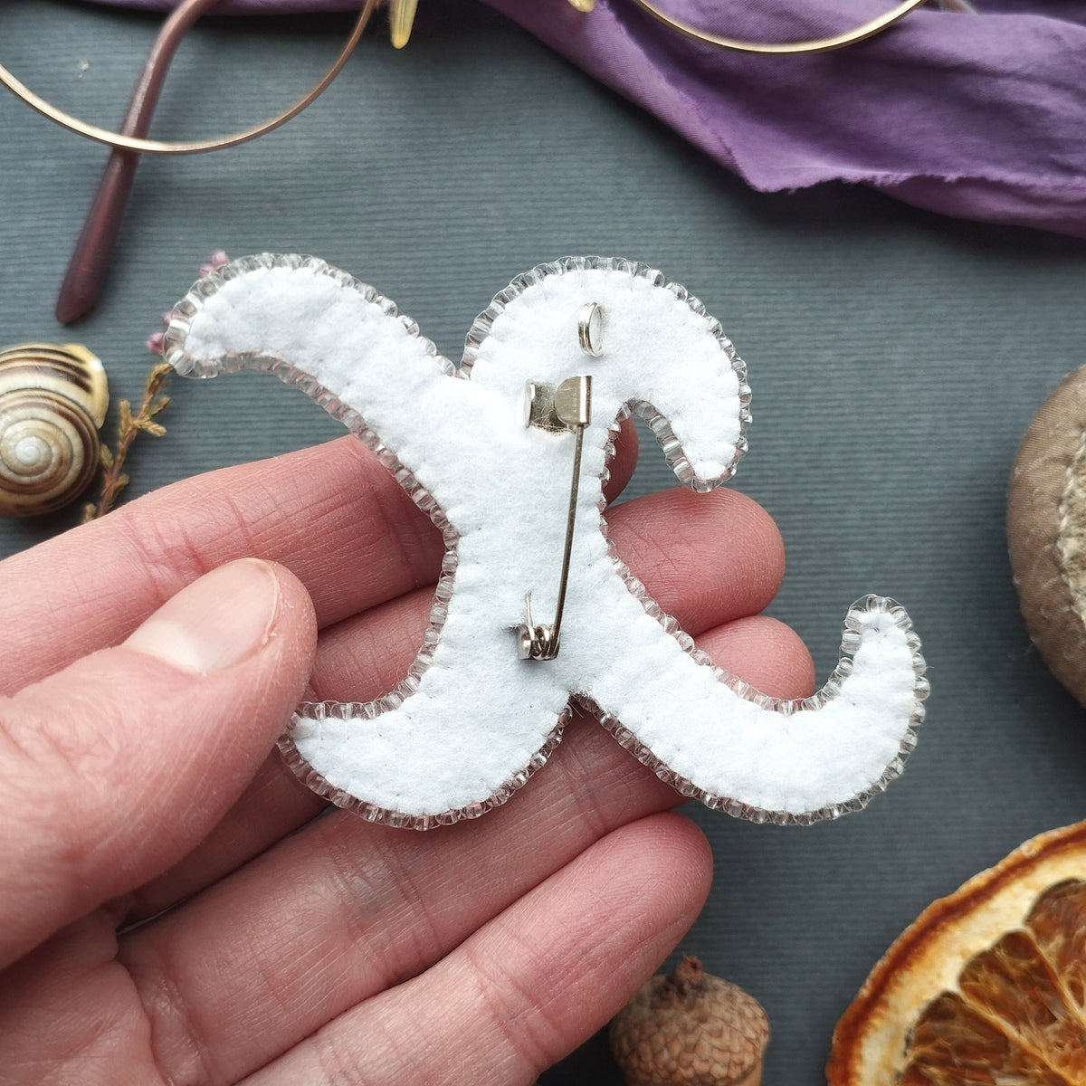 A hand holding a white fabric pin with a decorative design, surrounded by various objects such as a pair of glasses, a purple fabric, and dried fruit.