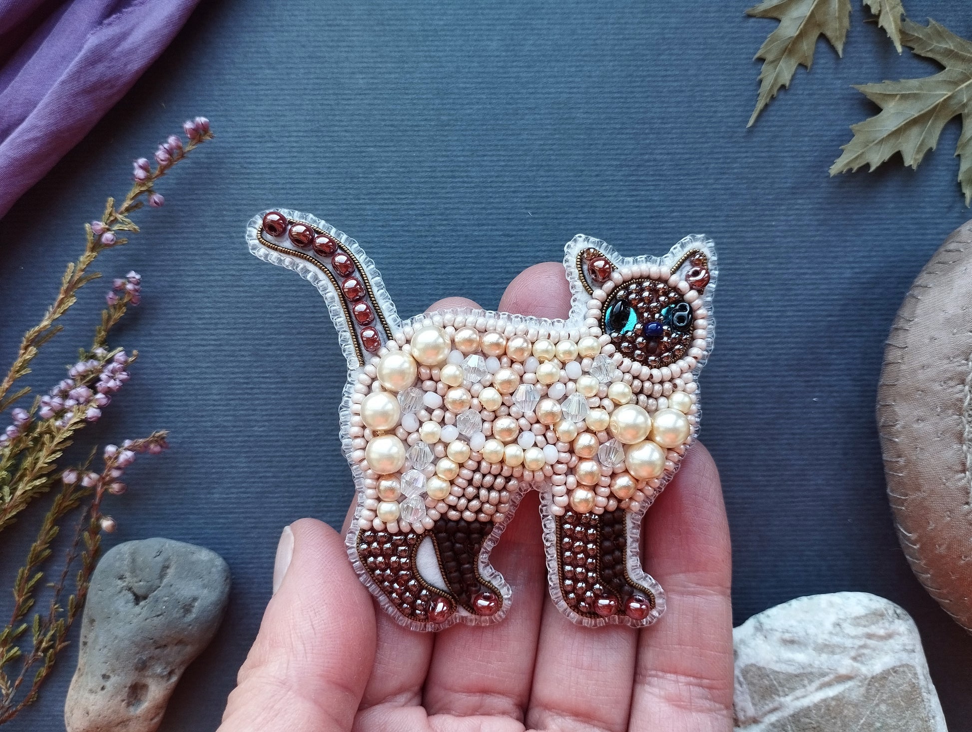 A hand holding a decorative beaded cat brooch against a dark background with various natural elements, such as leaves and rocks.