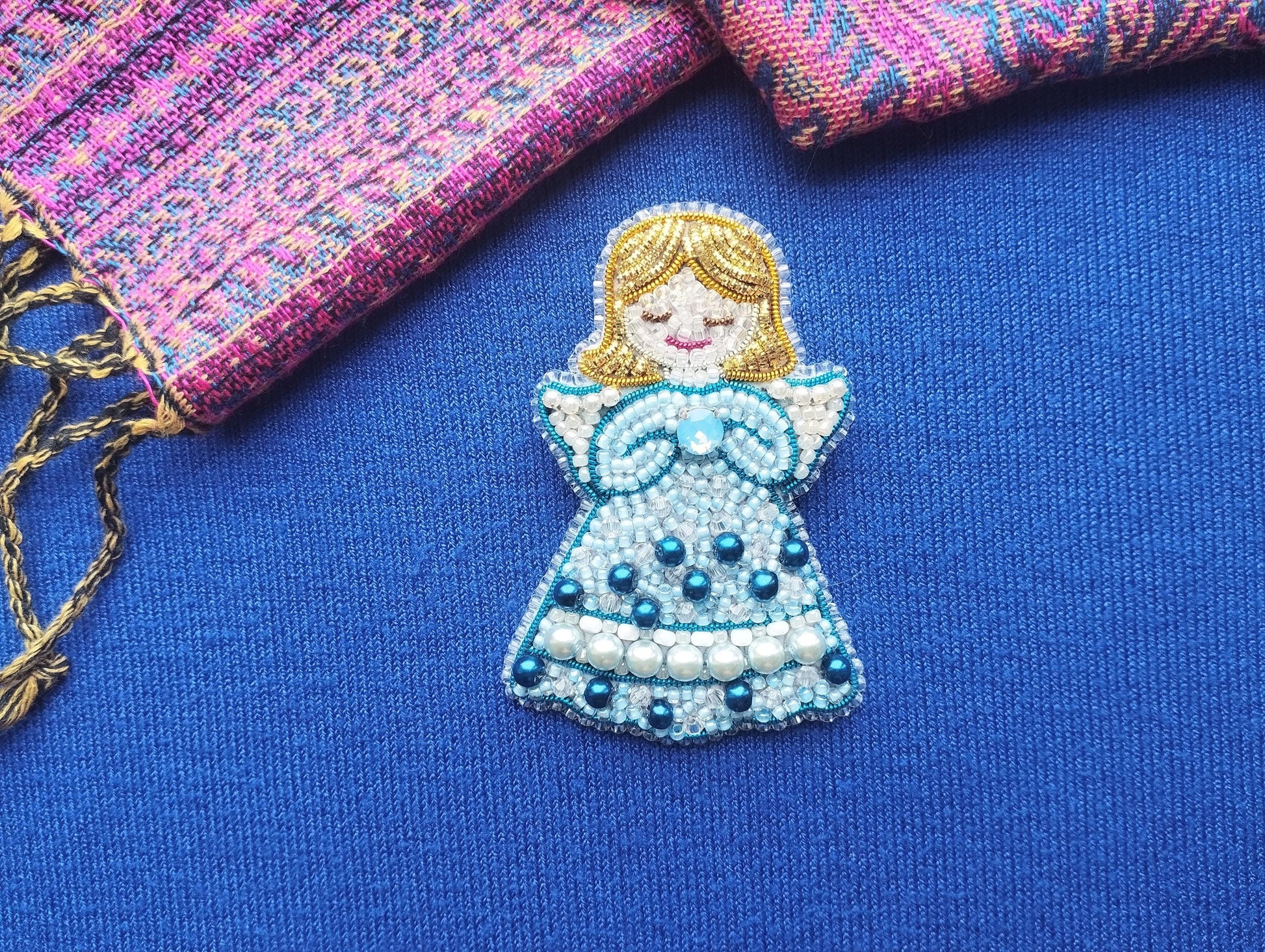 A hand-embroidered doll with a blue dress and white beads is displayed on a blue fabric background.