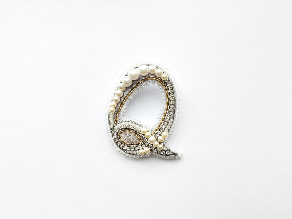 A decorative letter 'Q' made of pearls, diamonds, and other embellishments, displayed against a white background.