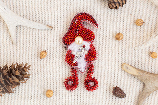 A red ornament resembling a gnome, adorned with beads and pearls, is the central focus of the image. Surrounding the ornament are various natural elements, including pine cones, antlers, and acorns, creating a festive and seasonal atmosphere.