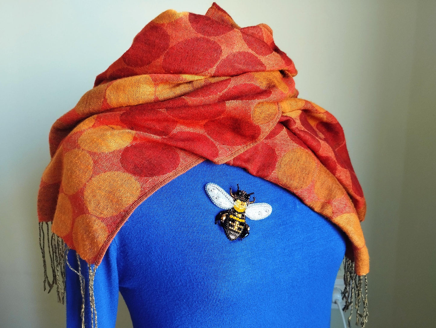 A woman wearing a blue sweater with a red and orange polka dot scarf draped over her shoulders, and a decorative bee brooch pinned to her sweater.