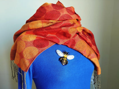 A woman wearing a blue sweater with a red and orange polka dot scarf draped over her shoulders, and a decorative bee brooch pinned to her sweater.