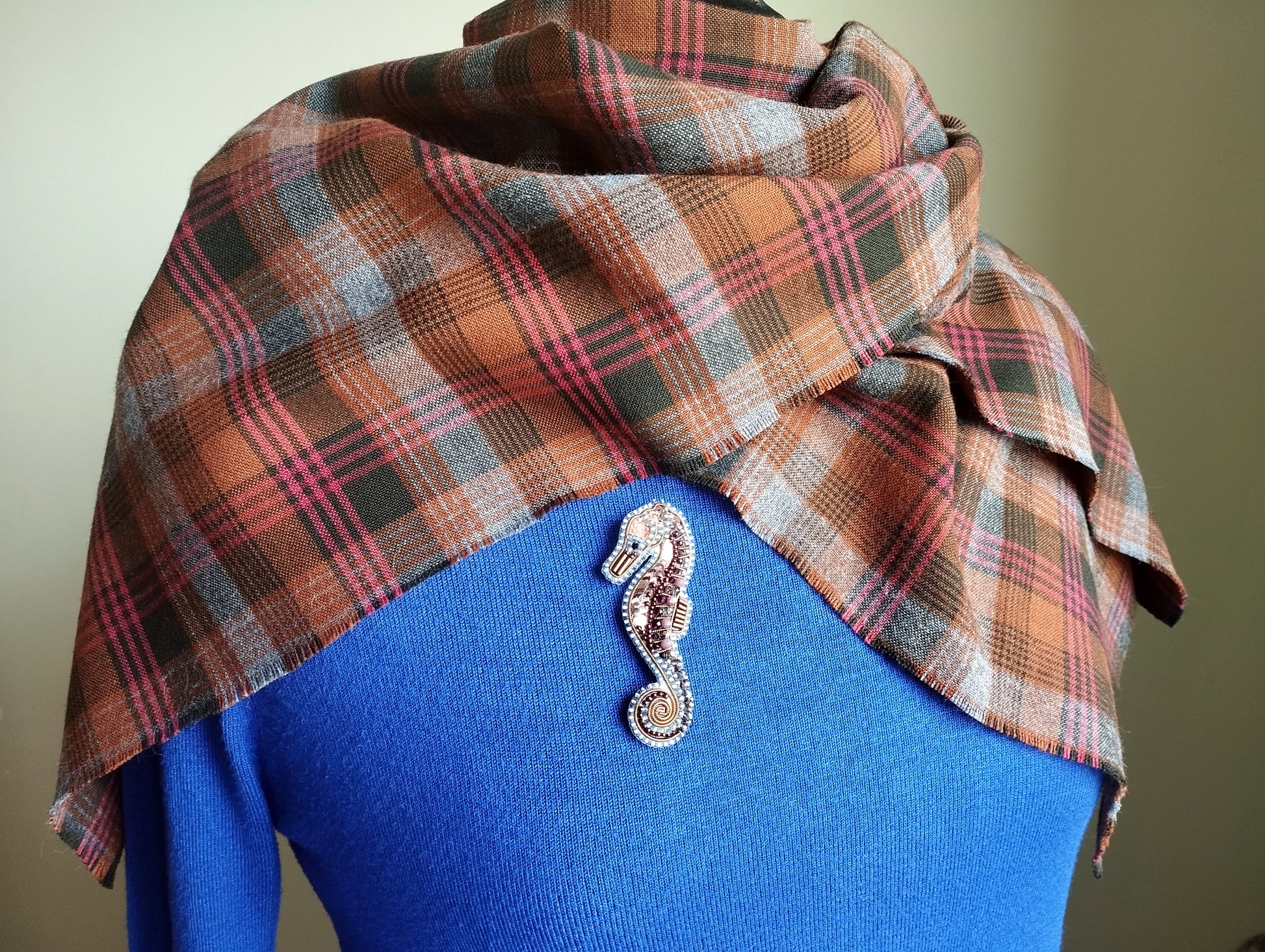 A plaid scarf is draped over a blue sweater, with a decorative seahorse brooch pinned to the sweater.