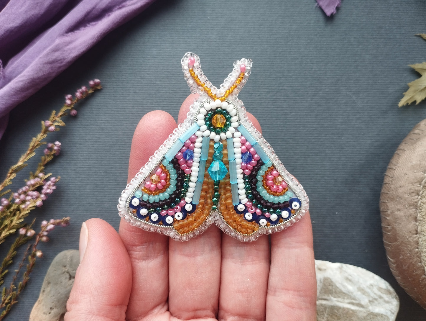 A hand holding a colorful, intricately beaded butterfly brooch against a dark background.