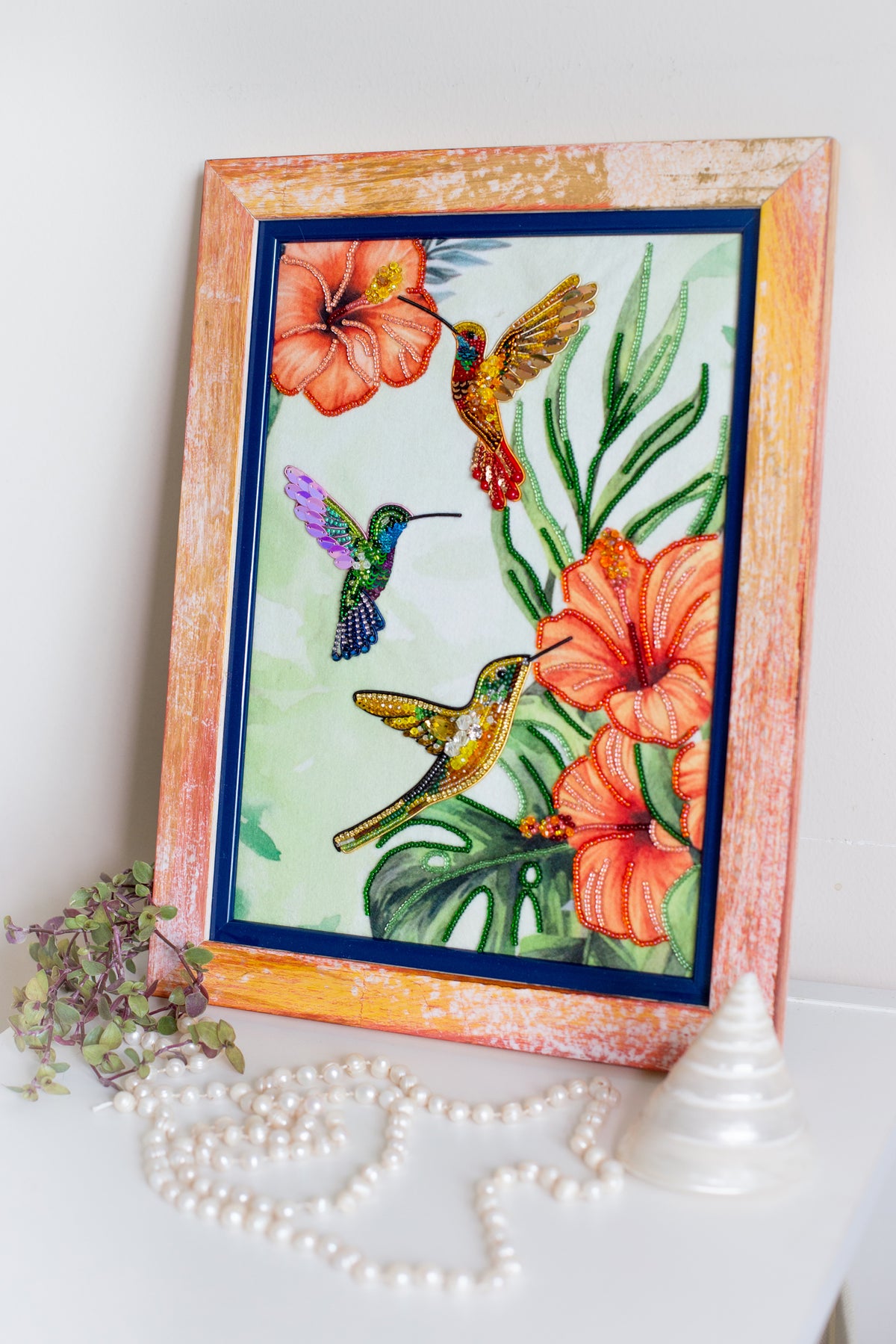 a framed painting of colorful hummingbirds surrounded by flowers, with a small white pyramid and a string of pearls on a white surface.