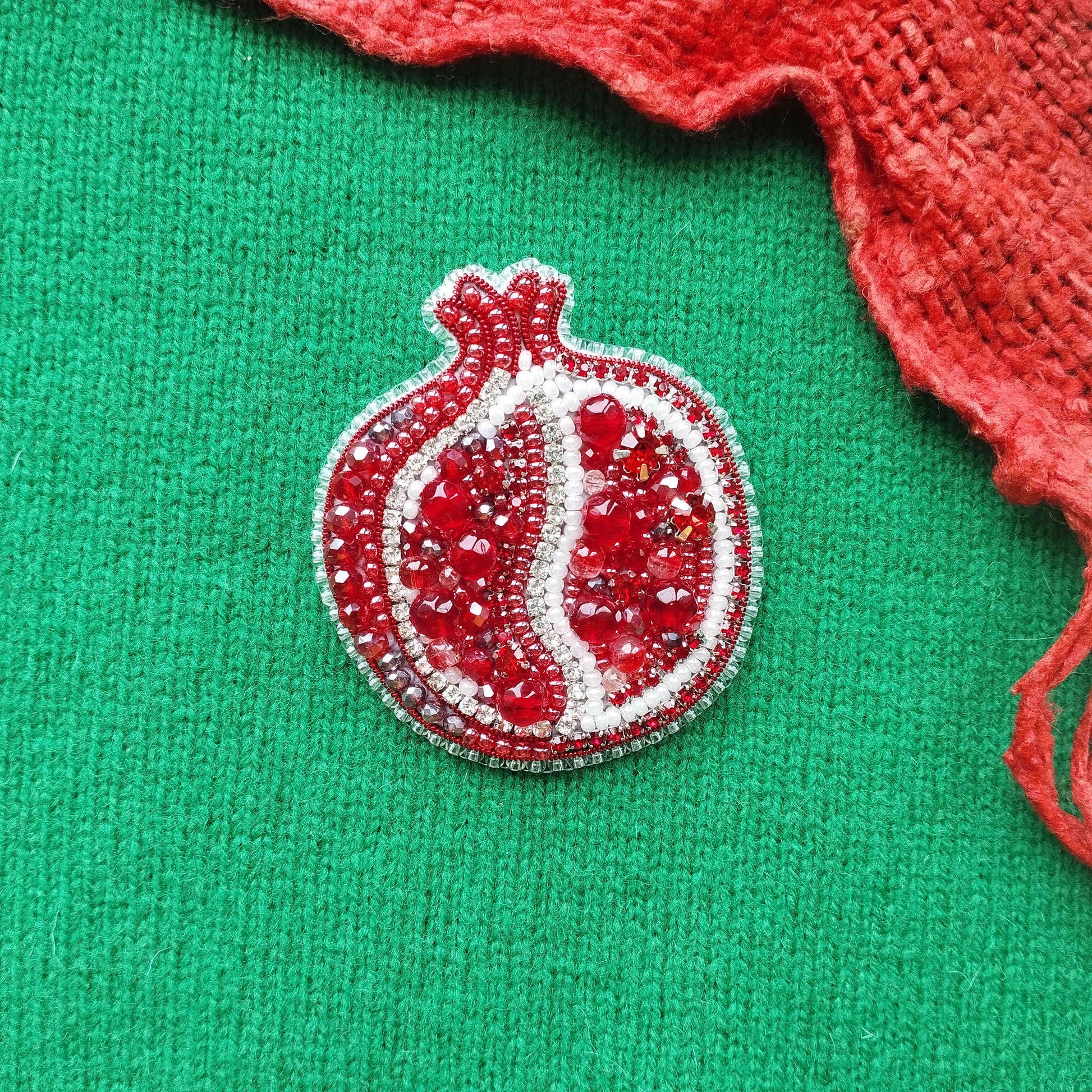 A red pomegranate-shaped brooch with red and white beads is displayed against a green background.