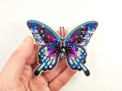 A hand is holding a colorful, intricately designed butterfly brooch made of sequins and beads.