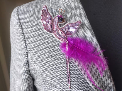 A close-up of a person wearing a gray tweed jacket with a decorative pin featuring a pink bird-like design and a feather attached to it.