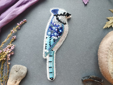 a detailed embroidered bird brooch with a blue and white color scheme, surrounded by various natural elements such as flowers, leaves, and rocks.