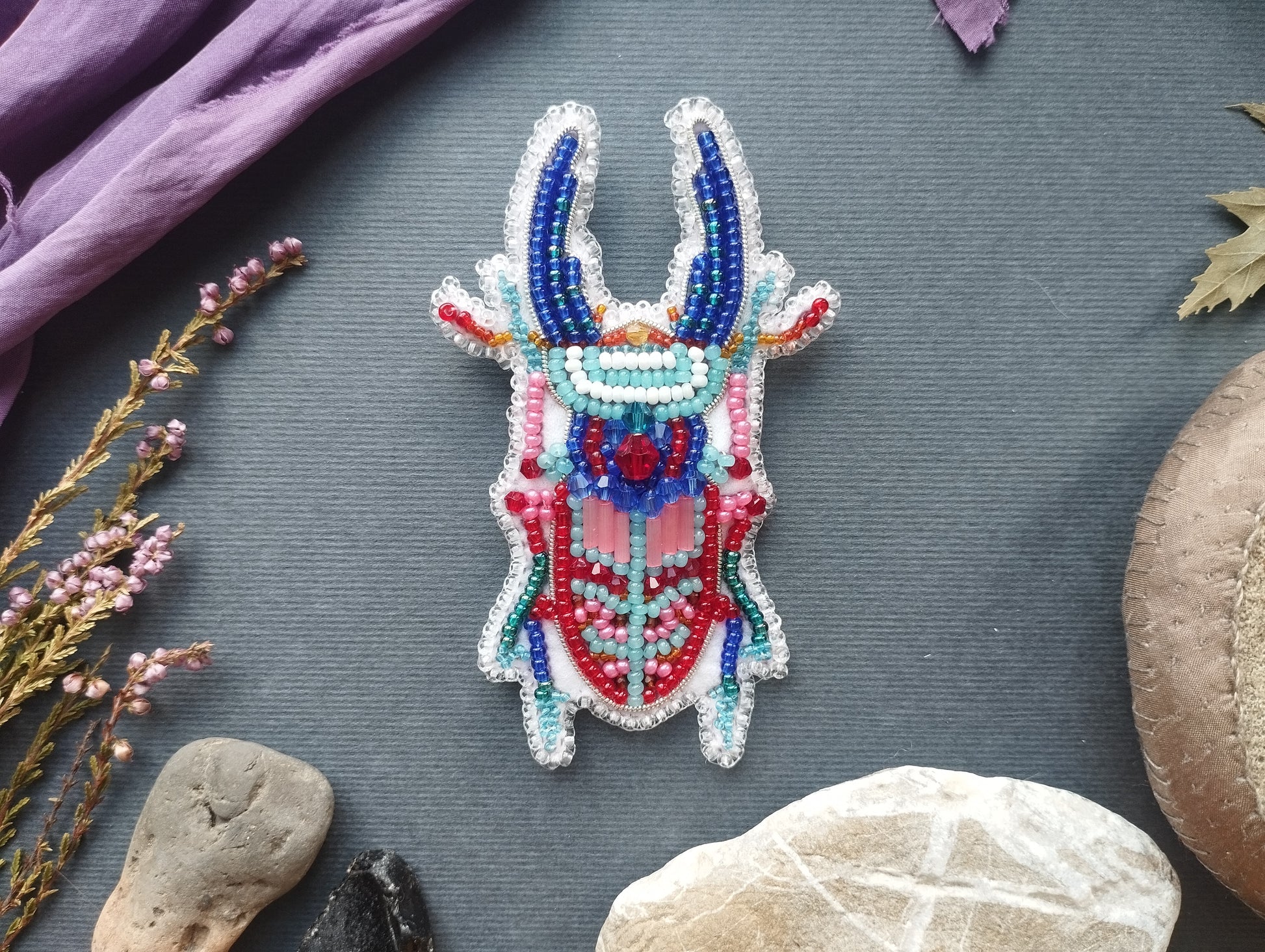 a colorful, intricately designed brooch or pin with a detailed, beaded pattern. The brooch is placed on a gray surface, surrounded by various objects such as rocks, leaves, and flowers.