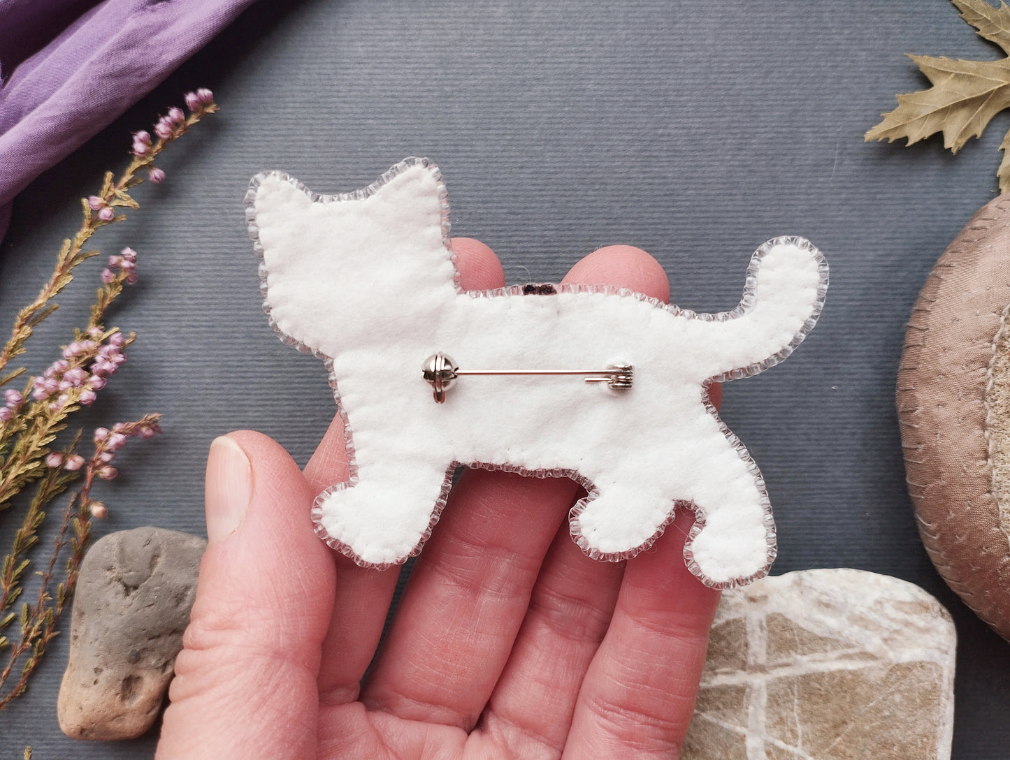 A hand holding a white felt cat pin against a gray background with various objects, including flowers and rocks.