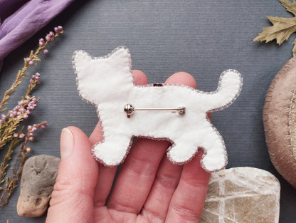 A hand holding a white felt cat pin against a gray background with various objects, including flowers and rocks.