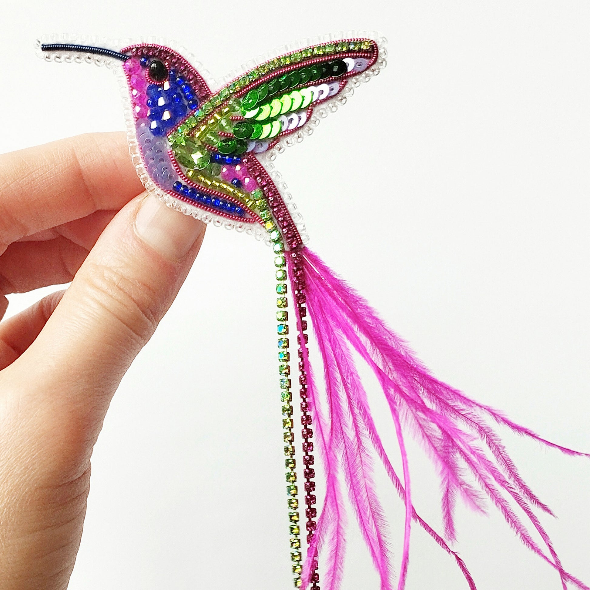 A hand is holding a colorful, intricately designed hummingbird brooch made of beads, feathers, and sequins.