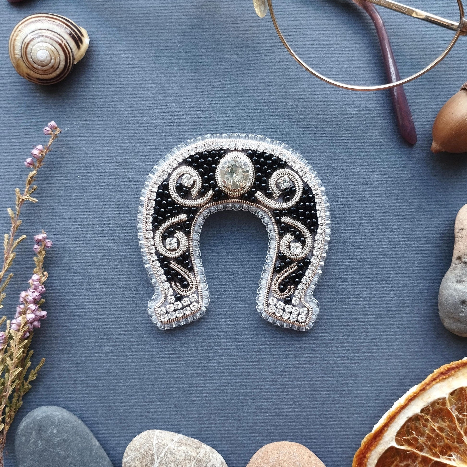 a horseshoe-shaped brooch adorned with jewels and beads, surrounded by various objects such as a magnifying glass, a seashell, a slice of orange, and a rock.