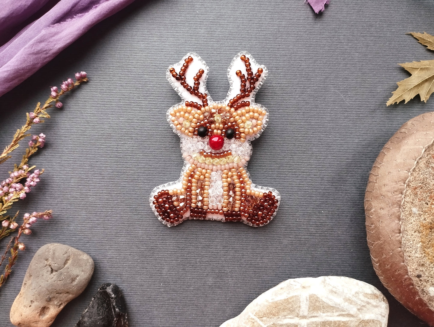 a beaded reindeer brooch with antlers, surrounded by various natural elements such as leaves, rocks, and a mushroom.