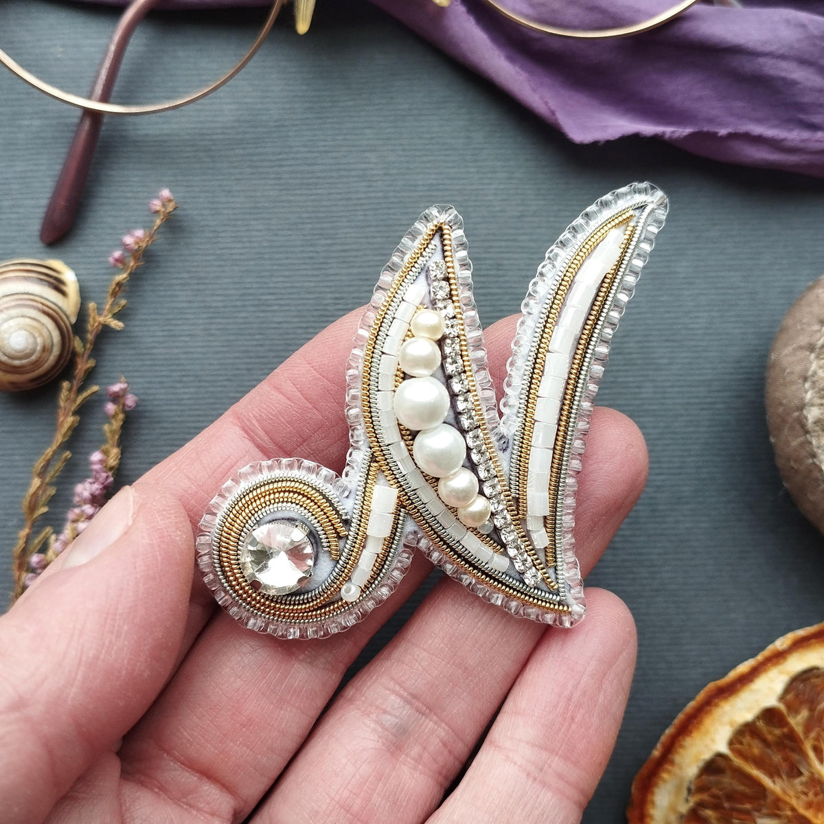 A hand holding a decorative piece of jewelry, which appears to be a brooch or pin, with various embellishments such as pearls and crystals.