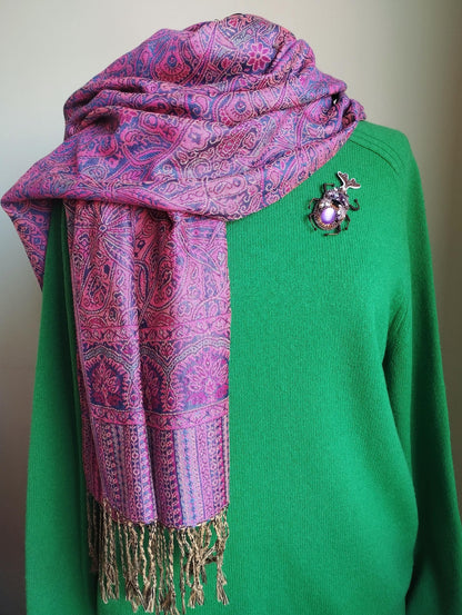 A mannequin is wearing a green sweater and a pink and purple patterned scarf with fringe, and a brooch is pinned to the sweater.