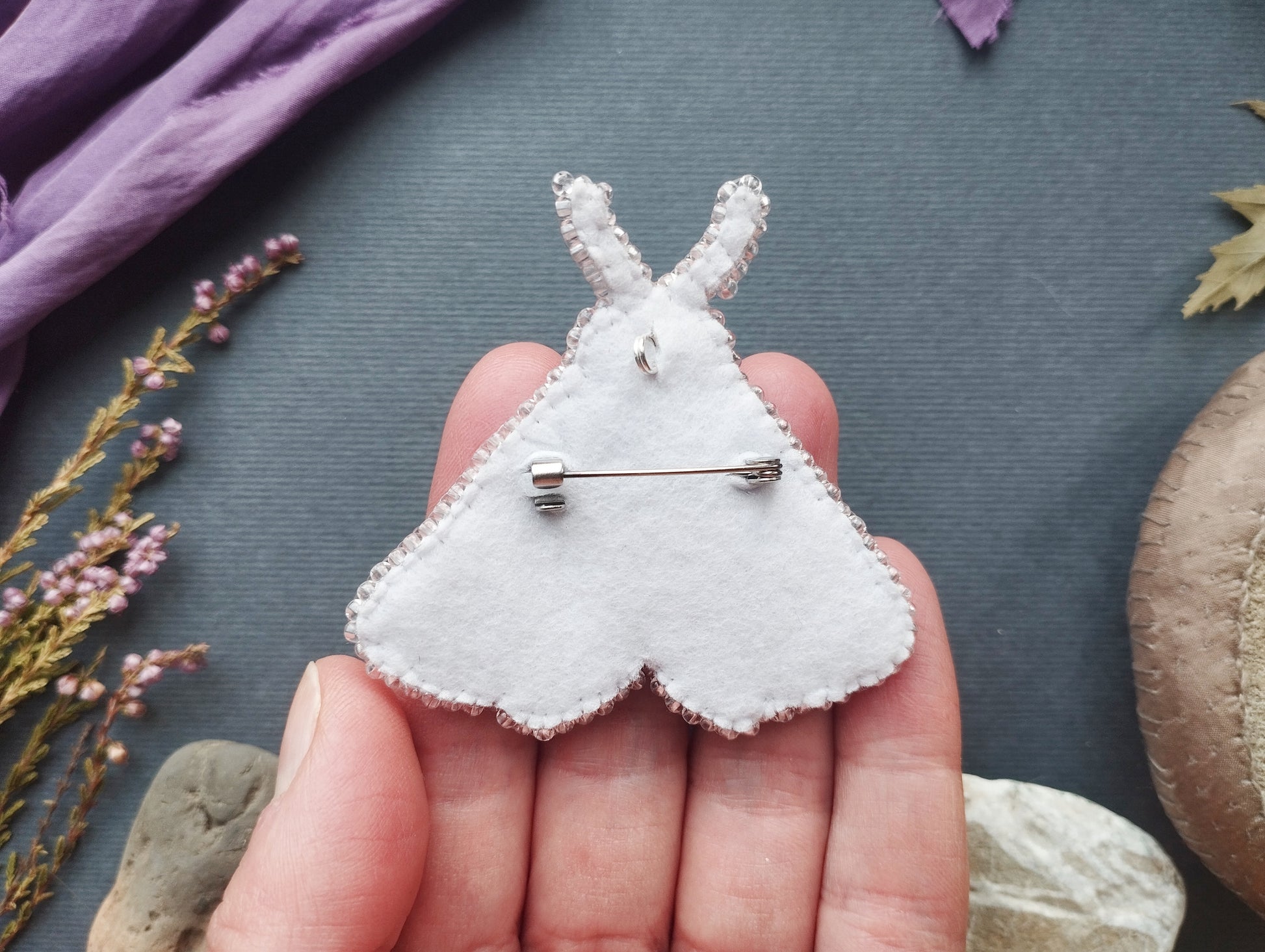 A hand holding a white felt butterfly pin against a dark background with some purple flowers and a brown object.