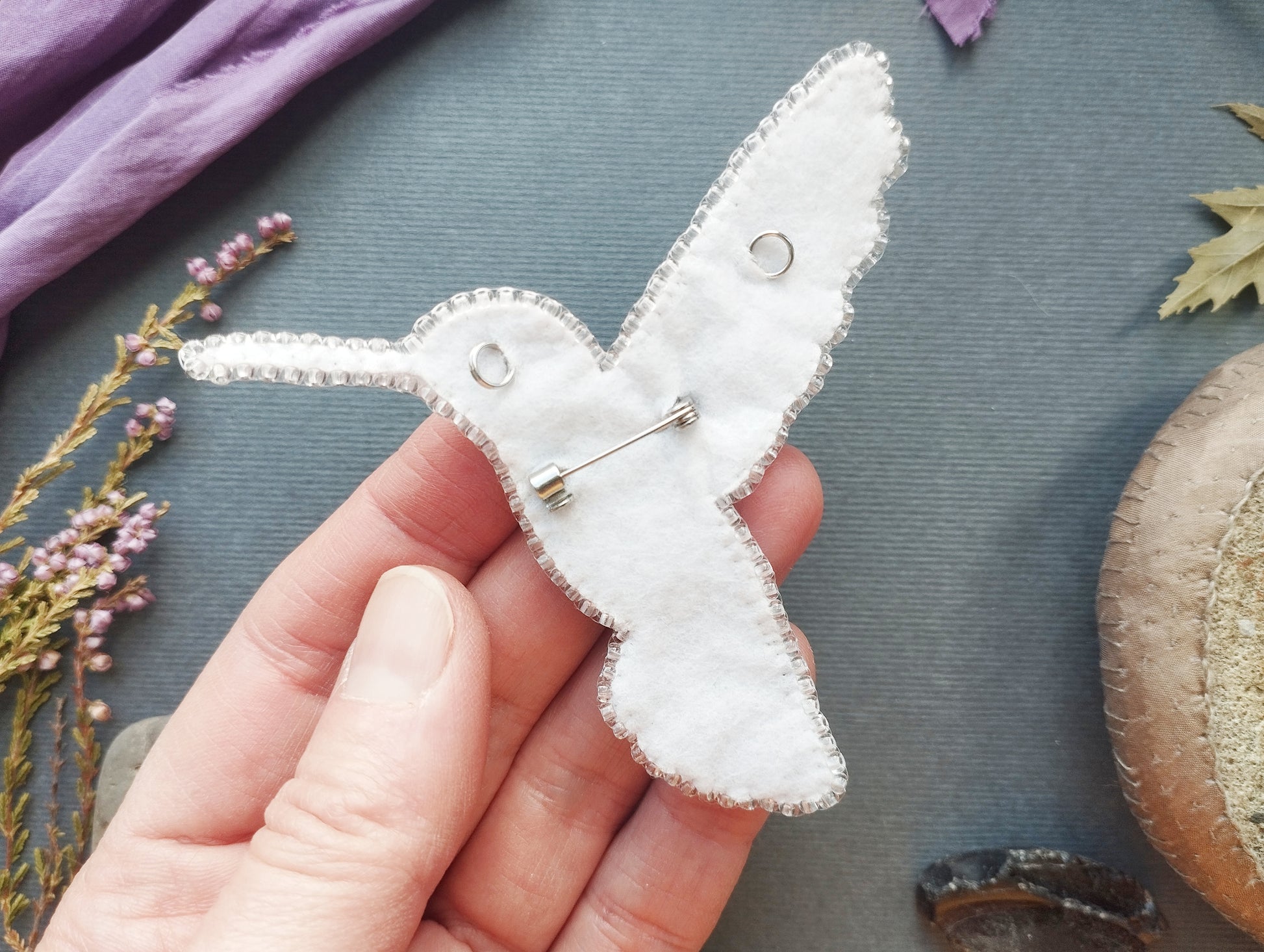 A hand is holding a white felt hummingbird pin against a blue background with various natural elements, such as flowers and leaves.