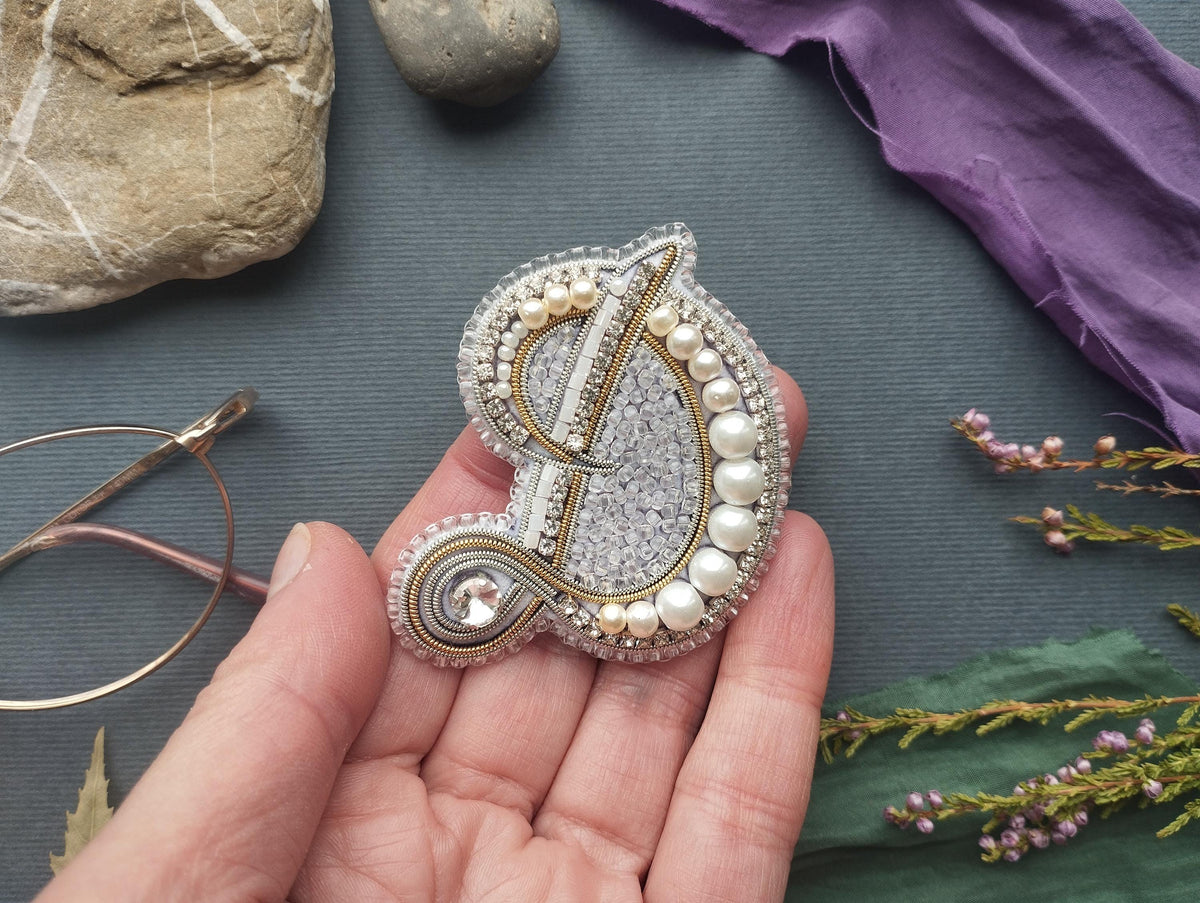 A hand holding a decorative button with pearls and a crystal element, surrounded by various objects such as a rock, a purple fabric, and some flowers.