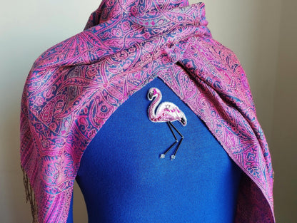 A mannequin wearing a blue top with a pink and purple paisley patterned scarf draped over it. The mannequin has a small, embroidered flamingo brooch pinned to the front of the blue top.