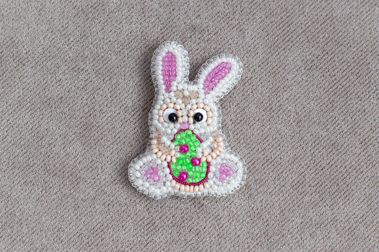 A beaded Easter bunny with a green egg in its mouth, sitting on a textured gray background.